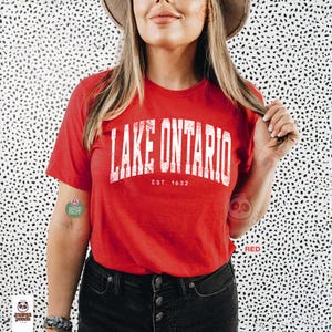 May include: A red short-sleeved t-shirt featuring the white distressed text LAKE ONTARIO arched across the chest with the smaller text EST. 1632 centered below. The shirt has a crew neckline and is worn with dark denim jeans and a tan hat.