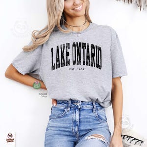 May include: A heather gray short-sleeve t-shirt featuring the black distressed text LAKE ONTARIO and EST. 1632 printed across the chest. The shirt has a crew neckline and is paired with blue denim jeans that have a distressed detail on the thigh.