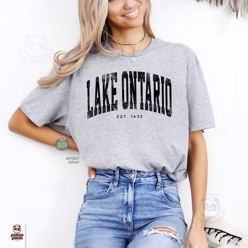 May include: A heather gray short-sleeve t-shirt featuring the black distressed text LAKE ONTARIO and EST. 1632 printed across the chest. The shirt has a crew neckline and is paired with blue denim jeans that have a distressed detail on the thigh.