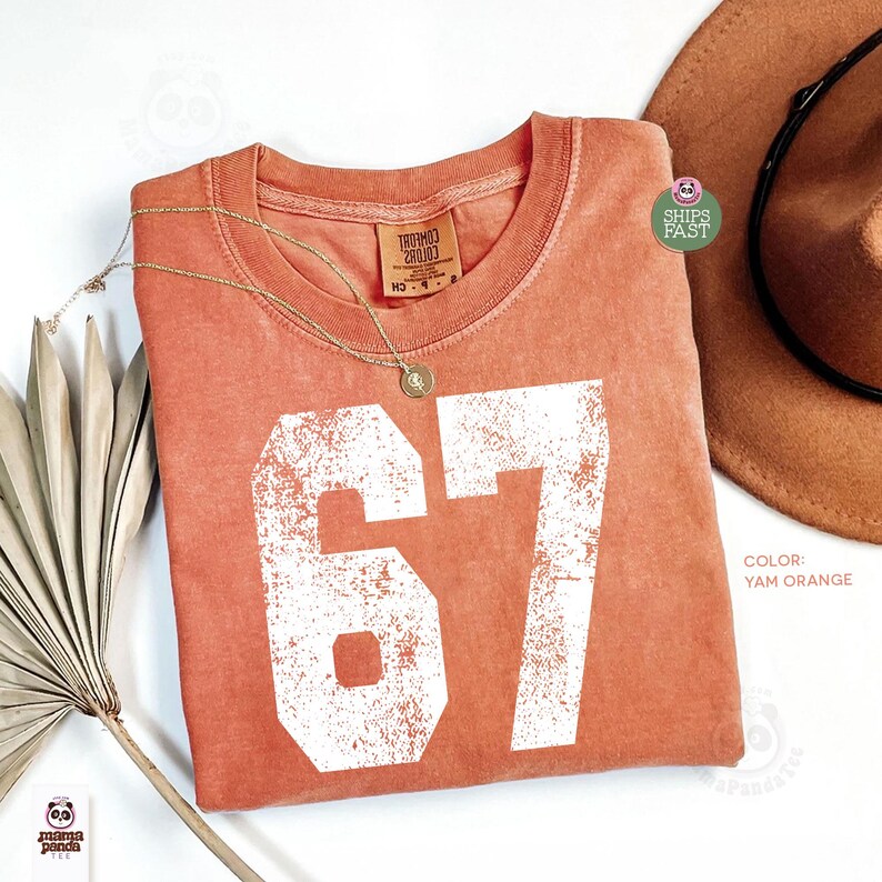 May include: A yam orange t-shirt with the number 67 in white, a gold necklace, and a brown hat. The shirt has a vintage, distressed look. The tag reads "THROUGH CLOUDS".