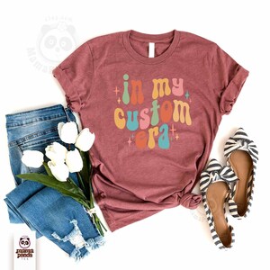 May include: A maroon t-shirt with the phrase "in my custom era" in colorful retro font. The shirt is styled with blue jeans, white tulips, and striped bow flats. The image is a flat lay.