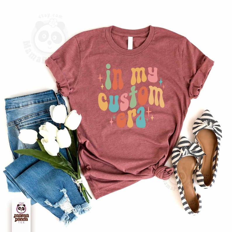 May include: A maroon t-shirt with the phrase "in my custom era" in colorful retro font. The shirt is styled with blue jeans, white tulips, and striped bow flats. The image is a flat lay.