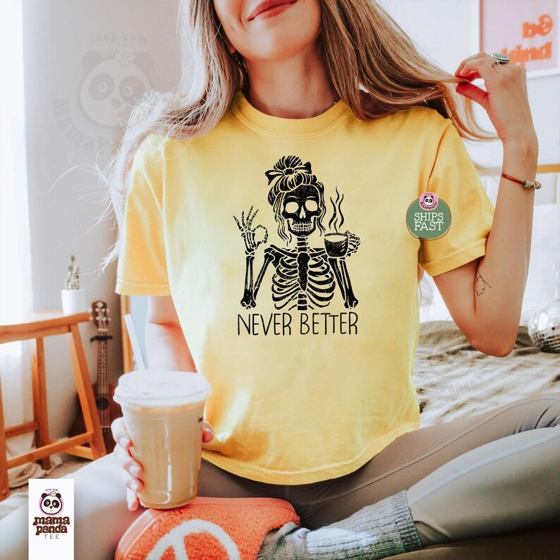 May include: A bright yellow short-sleeve t-shirt featuring a black graphic of a skeleton with a messy bun hairstyle, holding a coffee cup in one hand and making an okay gesture with the other, with the text NEVER BETTER printed below.