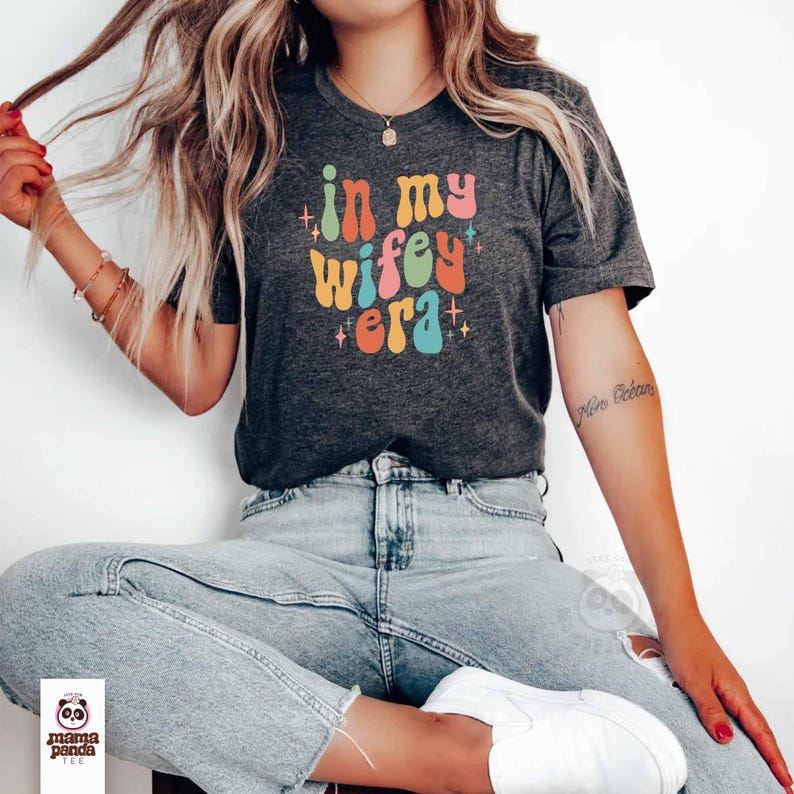 May include: Dark gray t-shirt with the text "in my wifey era" in colorful, retro-style lettering. The shirt features a crew neck and short sleeves. The brand logo "mama panda tee" is visible at the bottom left.