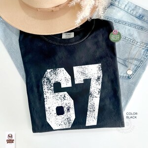 May include: Black t-shirt with the number 67 in distressed white lettering. The shirt is folded and placed on top of light-wash denim jeans. A tan hat and a silver necklace are also visible.