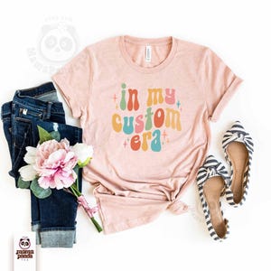 May include: A light pink t-shirt with the text "in my custom era" in colorful retro font. The shirt is styled with dark wash jeans, a bouquet of pink and white flowers, and striped bow flats.