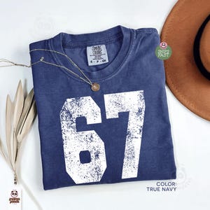 May include: A navy blue t-shirt with the number "67" in distressed white lettering. The shirt has a crew neck and a small tag at the neckline. A gold necklace with a pendant rests on the shirt. A brown hat is visible in the background.