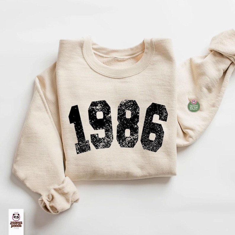 May include: Cream-colored sweatshirt with the year "1986" printed in distressed black block letters. The sweatshirt has a crew neck and long sleeves, with a small pin on the sleeve. The "mama panda tee" logo is visible at the bottom left.