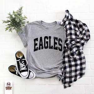 K&ouml;nnte beinhalten: Ein meliertes graues T-Shirt mit dem schwarzen Aufdruck "EAGLES", kombiniert mit schwarz-wei&szlig;en Turnschuhen und einem schwarz-wei&szlig; karierten Hemd. Eine kleine gr&uuml;ne Pflanze ist ebenfalls im Bild.
