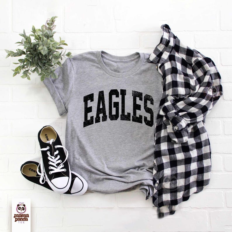 K&ouml;nnte beinhalten: Ein meliertes graues T-Shirt mit dem schwarzen Aufdruck "EAGLES", kombiniert mit schwarz-wei&szlig;en Turnschuhen und einem schwarz-wei&szlig; karierten Hemd. Eine kleine gr&uuml;ne Pflanze ist ebenfalls im Bild.