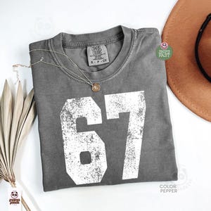 May include: A gray Comfort Colors t-shirt with the number 67 in white, featuring a distressed design. The shirt has a crew neck and a small tag. A gold necklace and a brown hat are also visible.