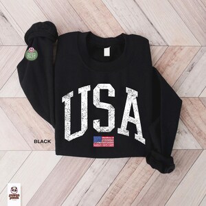 May include: Black crew neck sweatshirt with the word "USA" in distressed white lettering and a small American flag graphic. The sweatshirt is a long-sleeved top.