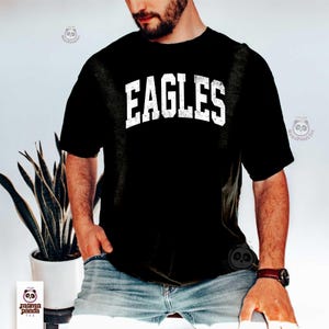 K&ouml;nnte beinhalten: Schwarzes T-Shirt mit dem Wort "EAGLES" in verblichenen wei&szlig;en Buchstaben. Das Shirt hat kurze &Auml;rmel und scheint aus einem weichen Material zu sein. Die Person, die das Shirt tr&auml;gt, tr&auml;gt auch eine blaue Jeans und eine Uhr.