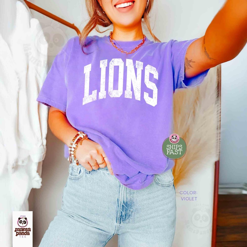 May include: A violet-colored t-shirt with the word "LIONS" printed in white block letters. The shirt is paired with light blue jeans. The image also includes a gold necklace and bracelet. The text "SHIPS FAST" is visible on the shirt.