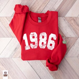 May include: A red sweatshirt with the year "1986" printed in large, distressed white letters. The sweatshirt is folded neatly, with the sleeves tucked in. The garment is made of a soft material, perfect for casual wear.