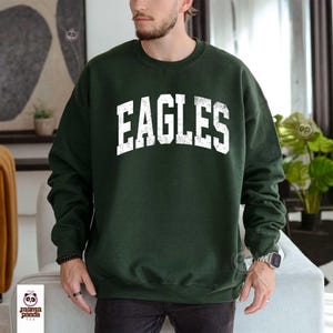 Może przedstawiać: Ciemnozielona bluza z nadrukiem "EAGLES" w dużych, białych literach na piersi. Bluza ma okrągły dekolt i długie rękawy. Zdjęcie przedstawia osobę noszącą bluzę.