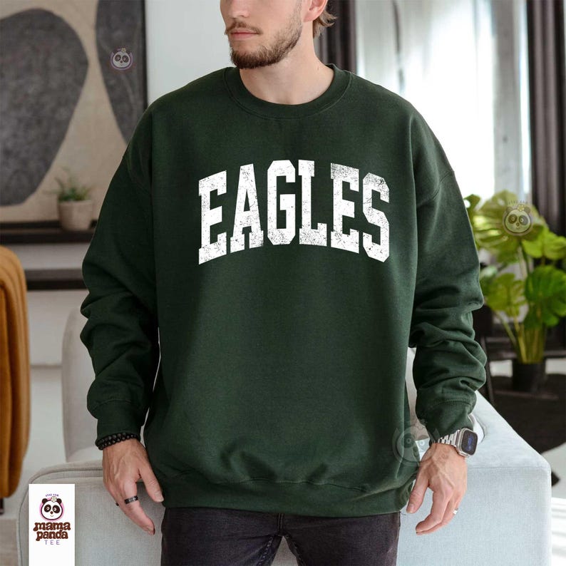 Może przedstawiać: Ciemnozielona bluza z nadrukiem "EAGLES" w dużych, białych literach na piersi. Bluza ma okrągły dekolt i długie rękawy. Zdjęcie przedstawia osobę noszącą bluzę.