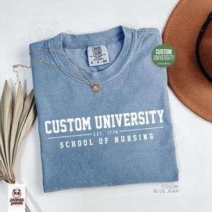 May include: A blue jean colored t-shirt with the text "CUSTOM UNIVERSITY SCHOOL OF NURSING" in white. A gold necklace and a brown hat are also visible. The shirt has a small tag that says "COMFORT COLORS".