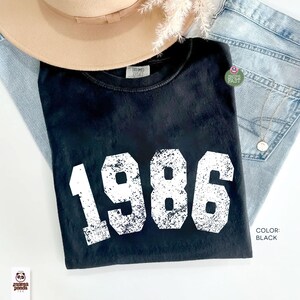 Może przedstawiać: Czarny t-shirt z rokiem 1986 wydrukowanym na biało, czcionką w stylu vintage. Koszulka jest złożona na jasnoniebieskich dżinsach, z beżowym kapeluszem i srebrnym naszyjnikiem. Kolor koszulki to czarny.