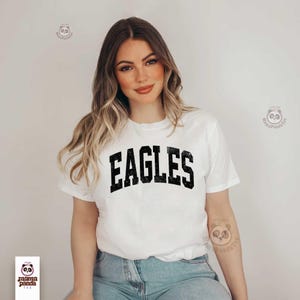 K&ouml;nnte beinhalten: Wei&szlig;es T-Shirt mit dem Wort "EAGLES" in verblichener schwarzer Schrift. Das T-Shirt hat einen Rundhalsausschnitt. Das Model tr&auml;gt eine blaue Jeans.