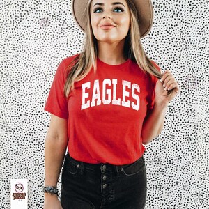 K&ouml;nnte beinhalten: Ein rotes T-Shirt mit dem Wort "EAGLES" in wei&szlig;en Blockbuchstaben. Das Shirt hat kurze &Auml;rmel und einen Rundhalsausschnitt. Die Person tr&auml;gt eine schwarze Jeans und einen braunen Hut.