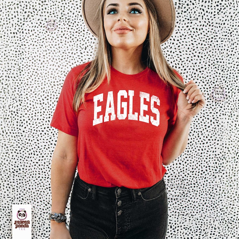 K&ouml;nnte beinhalten: Ein rotes T-Shirt mit dem Wort "EAGLES" in wei&szlig;en Blockbuchstaben. Das Shirt hat kurze &Auml;rmel und einen Rundhalsausschnitt. Die Person tr&auml;gt eine schwarze Jeans und einen braunen Hut.