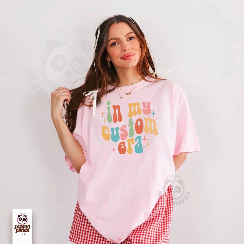 May include: A light pink t-shirt with the text "in my custom era" in colorful retro font. The shirt is paired with red and white checkered shorts. The Mama Panda Tee logo is visible in the bottom left corner.