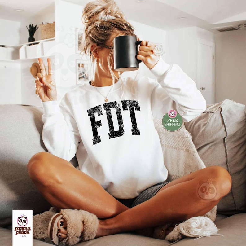 May include: White sweatshirt with the text "FDT" in black block letters. The letters have a distressed look. The sweatshirt is being worn by a person sitting on a couch.