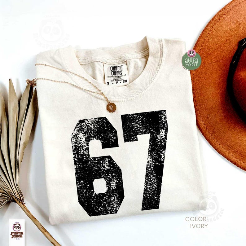 May include: Ivory-colored t-shirt with the number 67 in distressed black print. The shirt has a crew neck and a tag that reads "Comfort Colors". A gold necklace with a pendant is draped on the shirt. A brown hat is visible in the background.