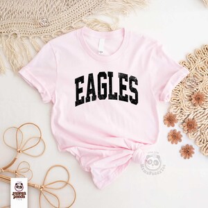 K&ouml;nnte beinhalten: Ein hellrosa T-Shirt mit dem Wort "EAGLES" in schwarzen Blockbuchstaben mit Used-Look. Das Shirt hat kurze &Auml;rmel und einen Rundhalsausschnitt. Das Shirt ist unten geknotet. Das Shirt befindet sich auf einer wei&szlig;en Oberfl&auml;che mit dekorativen Elementen.