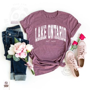 May include: A heather maroon short-sleeve t-shirt with the white text LAKE ONTARIO EST. 1632 printed on the front, paired with dark blue denim jeans, a bouquet of pink and white flowers, and a pair of light pink sneakers on a white background.
