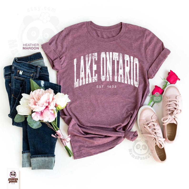 May include: A heather maroon short-sleeve t-shirt with the white text LAKE ONTARIO EST. 1632 printed on the front, paired with dark blue denim jeans, a bouquet of pink and white flowers, and a pair of light pink sneakers on a white background.