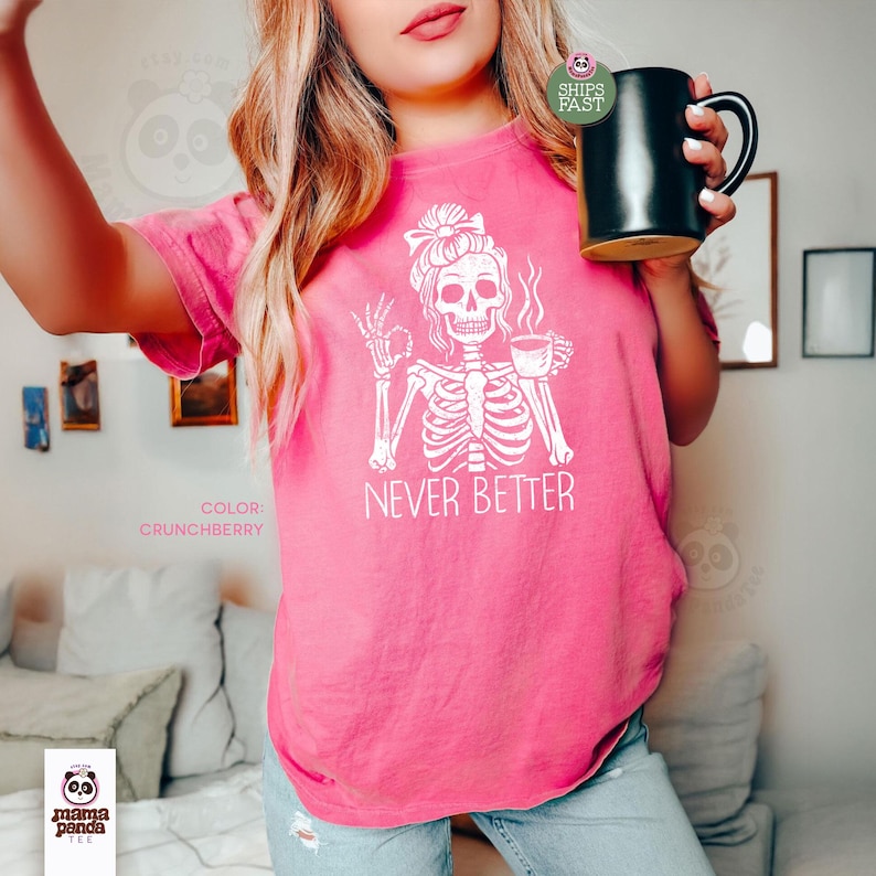 May include: A bright pink short-sleeve t-shirt featuring a white graphic of a skeleton with a messy bun holding a coffee cup. The skeleton is making an okay hand gesture. The text NEVER BETTER is printed in white below the graphic.