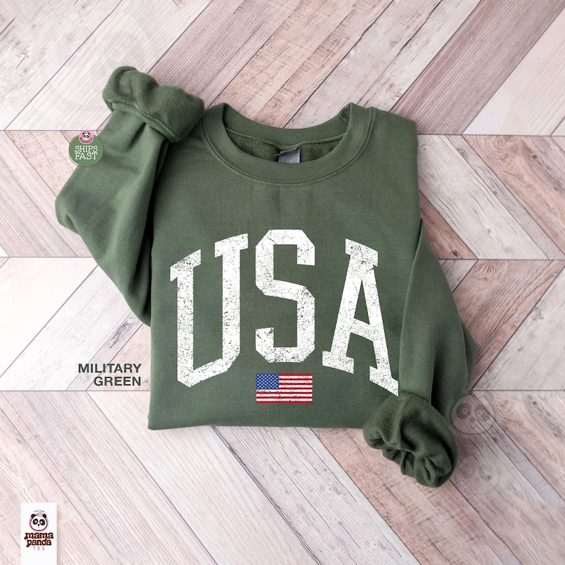 May include: Military green sweatshirt with the word "USA" in distressed white lettering and a small American flag graphic. The sweatshirt has a crew neck and long sleeves with ribbed cuffs. The garment is displayed on a light wood-grain surface.
