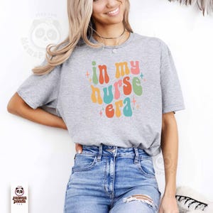 May include: A gray t-shirt with the text "in my nurse era" in colorful retro font. The design includes orange, pink, green, and blue letters with star accents. The shirt is paired with blue jeans.
