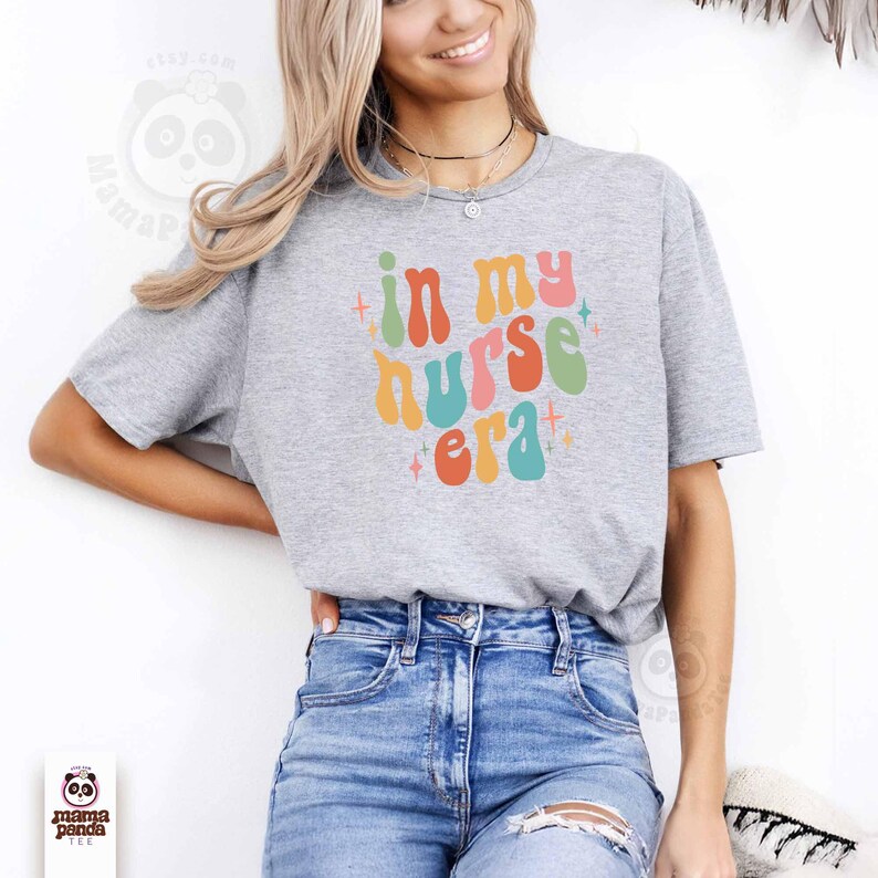 May include: A gray t-shirt with the text "in my nurse era" in colorful retro font. The design includes orange, pink, green, and blue letters with star accents. The shirt is paired with blue jeans.