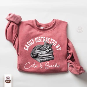May include: A pink sweatshirt with the text "Easily Distracted By Cats & Books" in white. A black and white illustration of a cat sleeping on a stack of books is above the text.