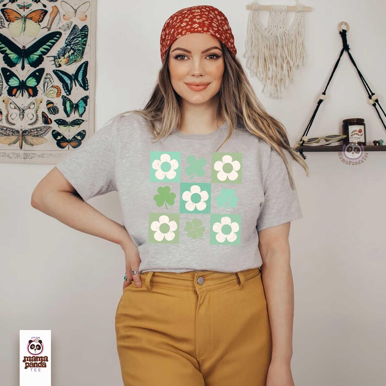Op de afbeelding: Een grijs T-shirt met een ontwerp van groene klavers en witte bloemen in een rasterpatroon. Het shirt wordt gedragen door een persoon met een rode bandana op het hoofd.
