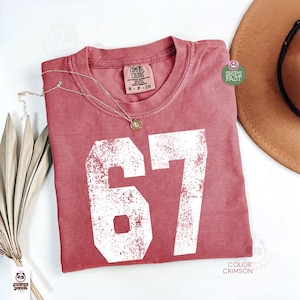May include: Crimson-colored t-shirt with the number 67 in white, featuring a distressed design. A gold necklace rests on the shirt. The shirt is labeled "Comfort Colors" and is size S-P-CH.
