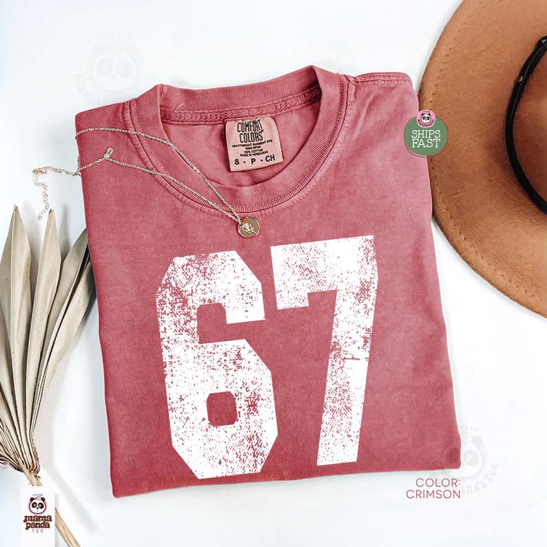 May include: Crimson-colored t-shirt with the number 67 in white, featuring a distressed design. A gold necklace rests on the shirt. The shirt is labeled "Comfort Colors" and is size S-P-CH.