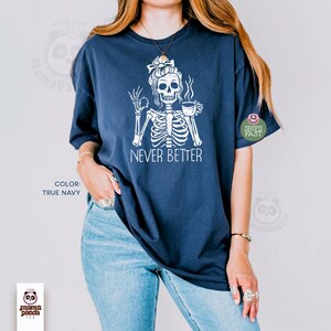 May include: A true navy short-sleeve t-shirt featuring a white graphic of a skeleton with a messy bun hairstyle, holding a coffee mug and making an okay hand gesture. The text NEVER BETTER is printed in white below the skeleton illustration.
