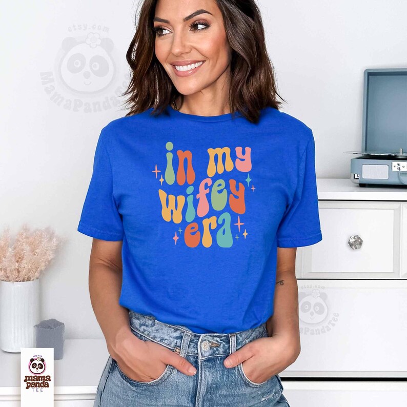 May include: A royal blue t-shirt with the text "in my wifey era" in colorful, retro-style lettering. The shirt is short-sleeved and worn by a person. The text is in orange, pink, yellow, and teal.