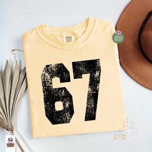 May include: A light yellow t-shirt with the number 67 printed in distressed black. The shirt is a Comfort Colors brand. The shirt is folded and has a round neck.