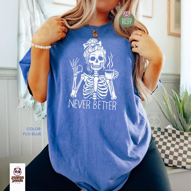 May include: Flo blue t-shirt with a white skeleton graphic. The skeleton is holding a coffee cup and making an "okay" hand gesture. The text "NEVER BETTER" is below the graphic. The shirt is a casual style.