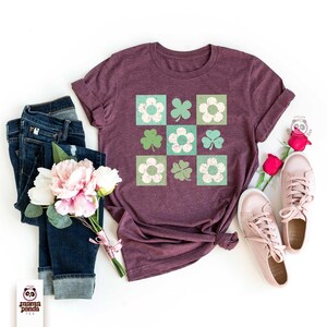 Op de afbeelding: Een maroon t-shirt met een grafisch ontwerp van een herhalend patroon van groene klavers en witte bloemen in vierkanten. Het shirt ligt plat op een wit oppervlak met een blauwe jeans, een boeket roze bloemen en een paar witte sneakers.