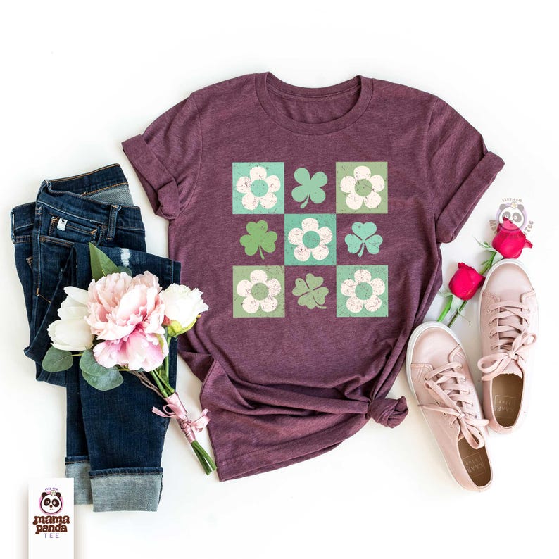Op de afbeelding: Een maroon t-shirt met een grafisch ontwerp van een herhalend patroon van groene klavers en witte bloemen in vierkanten. Het shirt ligt plat op een wit oppervlak met een blauwe jeans, een boeket roze bloemen en een paar witte sneakers.