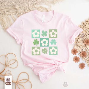 Op de afbeelding: Een roze T-shirt met een groen-wit klavertjevier en bloemenontwerp. Het ontwerp is in een geruit patroon.