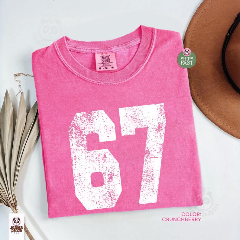 May include: A bright pink t-shirt with the number "67" printed in white. The shirt has a crew neck and short sleeves. The color is "Crunchberry". A brown hat is partially visible in the background.