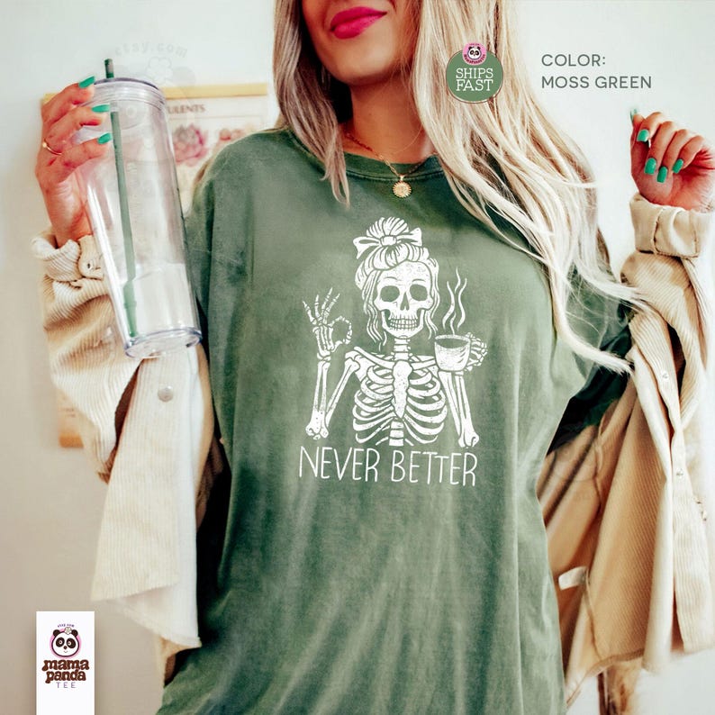 May include: A moss green t-shirt featuring a white graphic of a skeleton with a hair bow, holding a coffee cup and making an okay hand gesture. The text NEVER BETTER is printed below the skeleton. The shirt is worn with a light-colored jacket.