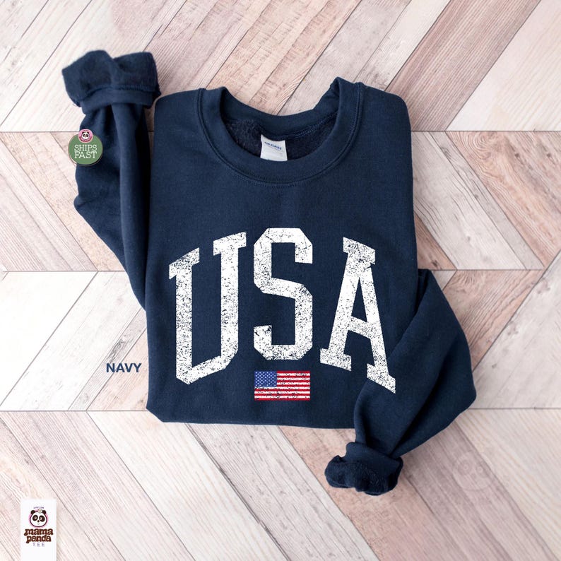 May include: Navy blue sweatshirt with the word "USA" in distressed white lettering. Below the text is a small American flag. The word "NAVY" is printed in white on the bottom left of the sweatshirt.